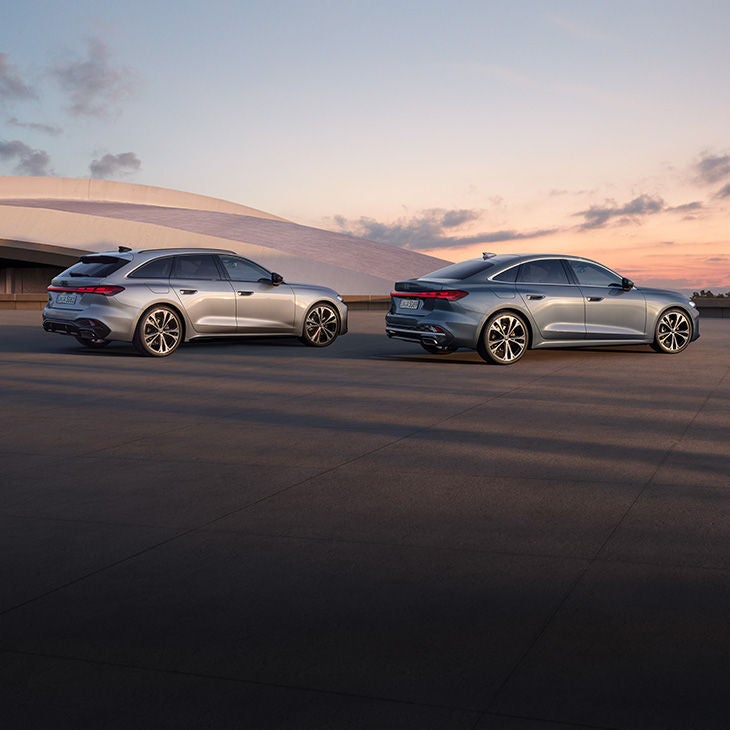  Zwei Audi-Fahrzeuge, ein grauer Kombi und eine graue Limousine, parken nebeneinander auf einer Straße. Im Hintergrund ist ein rosafarbener und orangefarbener Himmel zu sehen.